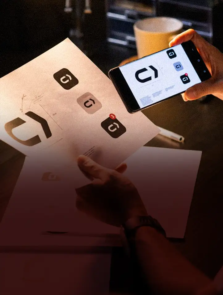Mãos segurando um celular com uma marca na tela, sobre papéis de identidade visual